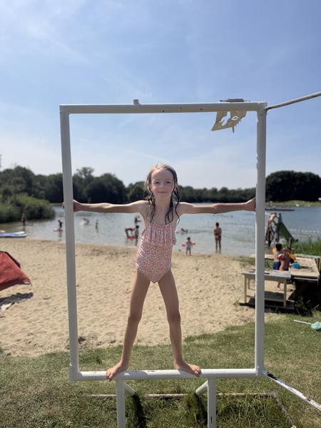 A young girl in a pink floral swimsuit playfully poses within a white frame on a sandy beach at Haarweg, Vuren